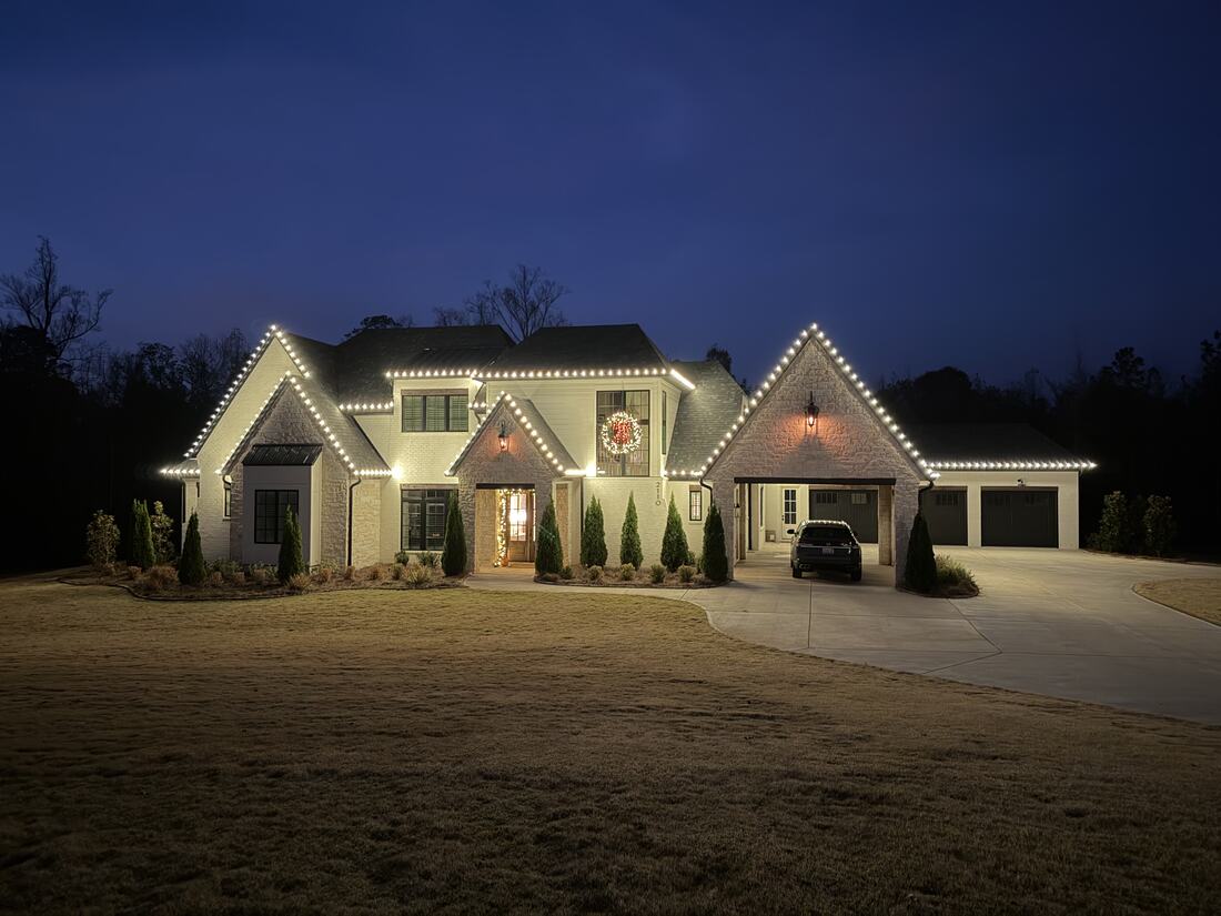A large cream brick home at dusk lined with warm white roofline lights and a lit wreath over the entry