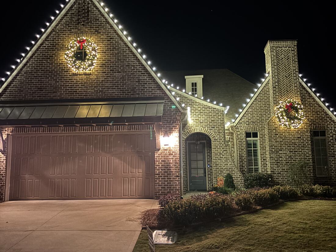 A brick home at night with lights along the gable and lit wreaths on the gable and chimney