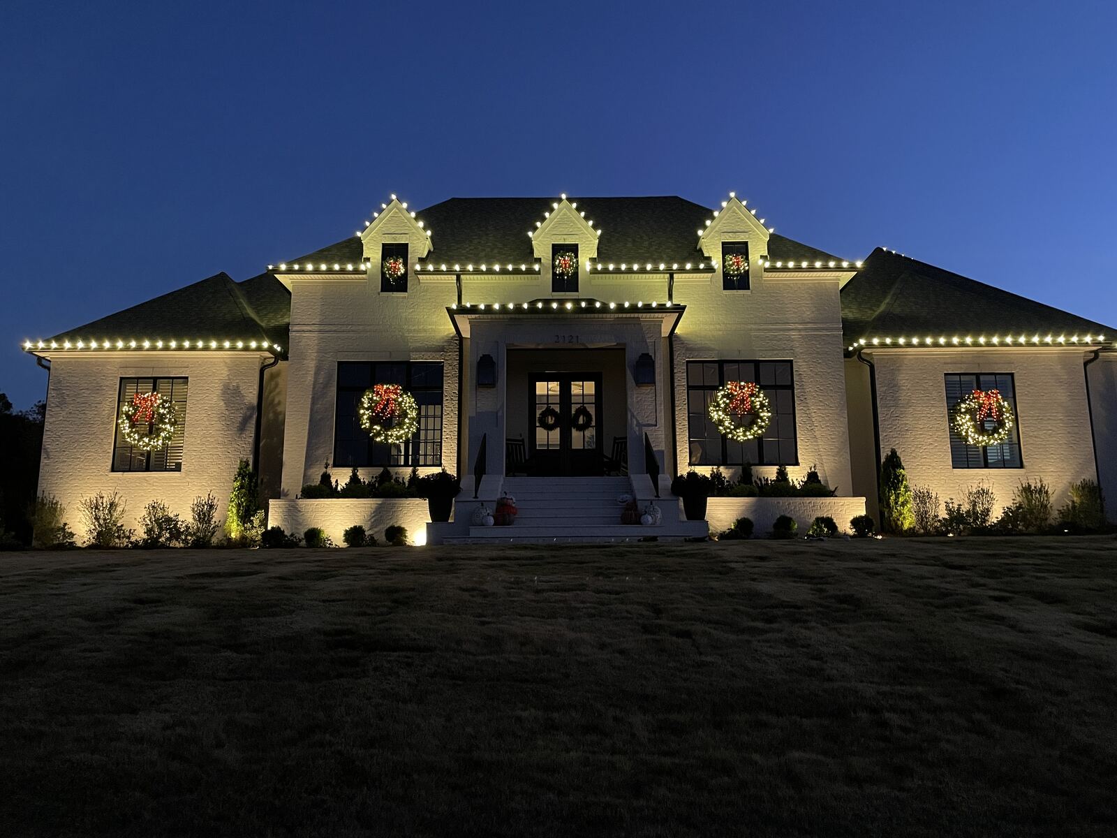 A large white brick home at dusk with warm white roofline lights and lit wreaths in every window